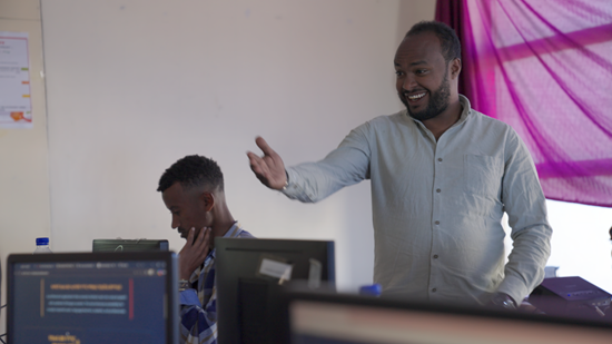 Man gestures and smiles at students at a computer bank 