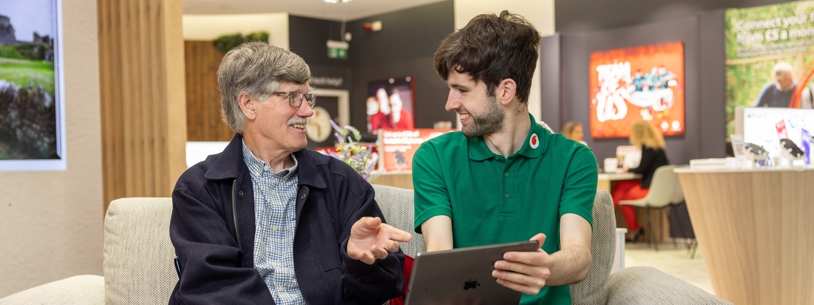 An older man sits with a Vodafone store assistant. The assistant helps the man with a tablet